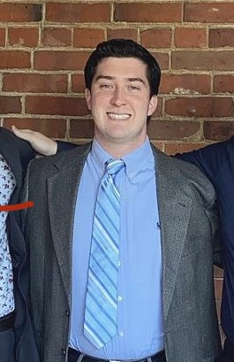 Christian Quigley, a white man with short black hair, poses against a brick wall. He's wearing a light blue dress shirt, a grey jacket, and a tie with diagonal stripes in different shades of blue and cream.