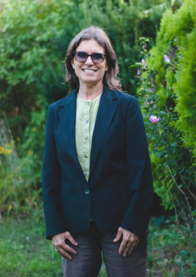 Image of a smiling woman wearing a suit jacket and sunglasses, in front of beautiful hedges
