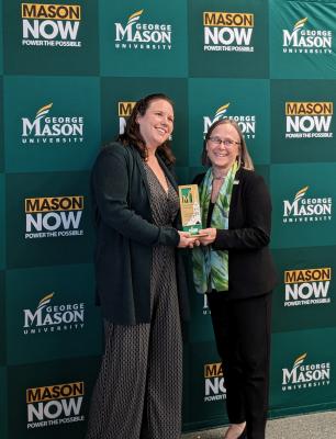 Laura Fox, tall with brown hair and jumpsuit, with Dean Ardis in black suit and scarf holding the award in front of a green wall with George Mason Now graphics