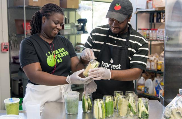 Two workers in a kitchen smiling and packing cucumbers into pickle jars.