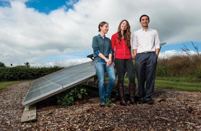 Three students stand in front of a solar panel in a field, smiling.