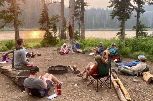 Students sitting in a circle at a campsite next to an alpine lake