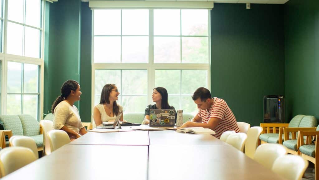 Students with laptops and books at desk sitting in front of tall windows.