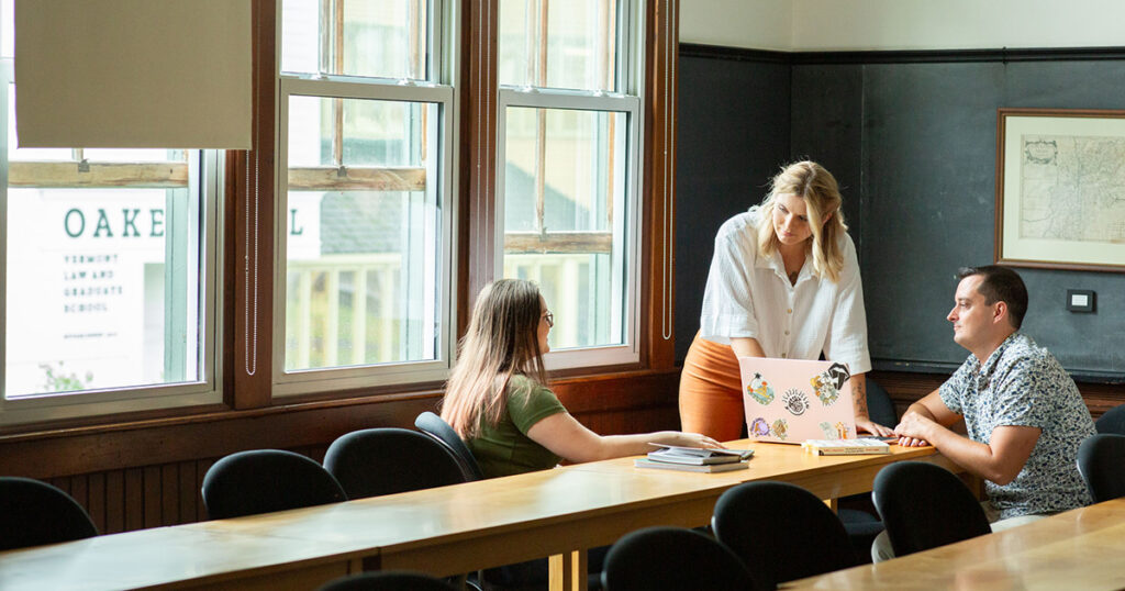 Three adults in a sunny classroom gather around a laptop.